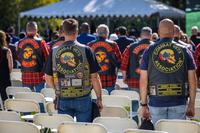 Facing away from the camera, male veterans stand amid folding chairs while wearing vests embroidered with military affiliations.