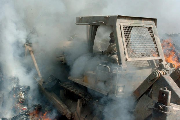 Smoke billows in from all sides as a bulldozer pushes into the flames of a burn pit in Iraq