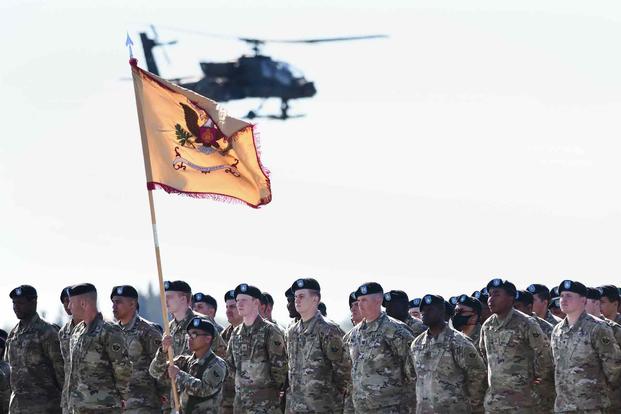 Soldiers stand in formation as an AH-64 Apache helicopter flies overhead.