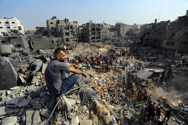 A man sits on the rubble as others wander among debris of buildings