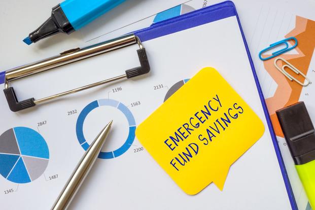 The words "emergency fund savings" appear on a yellow sticky note that's stuck, along with a silver pen, to a set of graphs on a clipboard next to a blue highlighter.