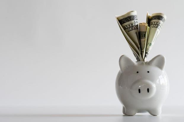 Folded cash sticks out of the top of a white piggy bank against a white background.