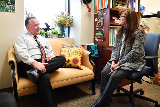 A man sits on a couch apparently talking to a woman sitting in a chair, in a posed picture in a cheerily lit office.