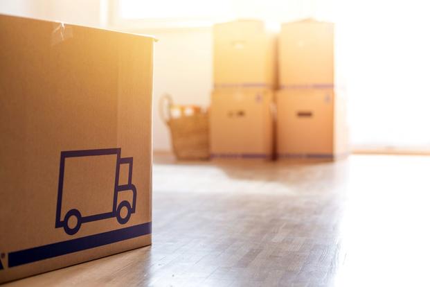 Sitting on a hardwood floor in a room with bright, diffused light, are moving boxes. An icon of a moving truck appears on the box in the foreground.