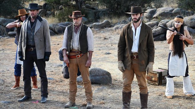 Three cowboys stand under guard in a rocky desert clearing as two Apache warriors hold them at gunpoint in the Western film “Frontier Crucible.”