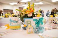 Gifts and bouquets in pastel blue and yellow are arranged on a table in a fluorescent-lit, institutional-type room.