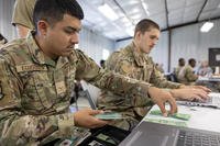 Air Force airmen in camouflage uniforms sit at a table in front of laptops, while one counts out fake money from a stack.