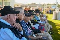 A row of older male military veterans, seated at a ceremony, mostly wear dark-colored ball caps signifying aspects of their military service.