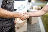 Two men's hands shake in the foreground in front of piles of moving boxes.