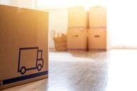 Sitting on a hardwood floor in a room with bright, diffused light, are moving boxes. An icon of a moving truck appears on the box in the foreground.