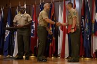 On a flag-draped stage, a Marine shakes another Marine's hand while a third Marine looks on.