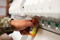 A hand holds a pill bottle under a dispenser.