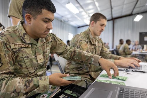 Air Force airmen in camouflage uniforms sit at a table in front of laptops, while one counts out fake money from a stack.